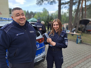 Umundurowana policjantka zajmująca się profilaktyką społeczną wspólnie z umundurowanym dzielnicowym stoją przed oznakowanym radiowozem.