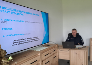 Na zdjęciu umundurowany policjant siedzi za biurkiem. Po lewej stronie widać tablicę interaktywną, na której widnieje slajd z opisem debaty społecznej.