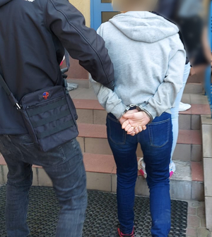 Policjant kryminalny w ubraniu cywilnym prowadzi kobietę, która ma założone kajdanki z tyłu.