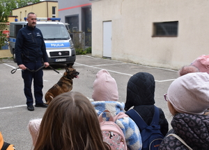 Umundurowany policjant przewodnik psa opowiada grupie dzieci na temat służby zwierząt w policji.