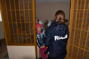 Umundurowana policjantka stoi w pomieszczeniu dla osób zatrzymanych  i opowiada dzieciom na temat bezpieczeństwa.