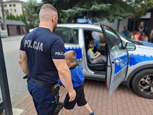 Umundurowany policjant stoi przy radiowozie oznakowanym, do którego wchodzą dzieci.